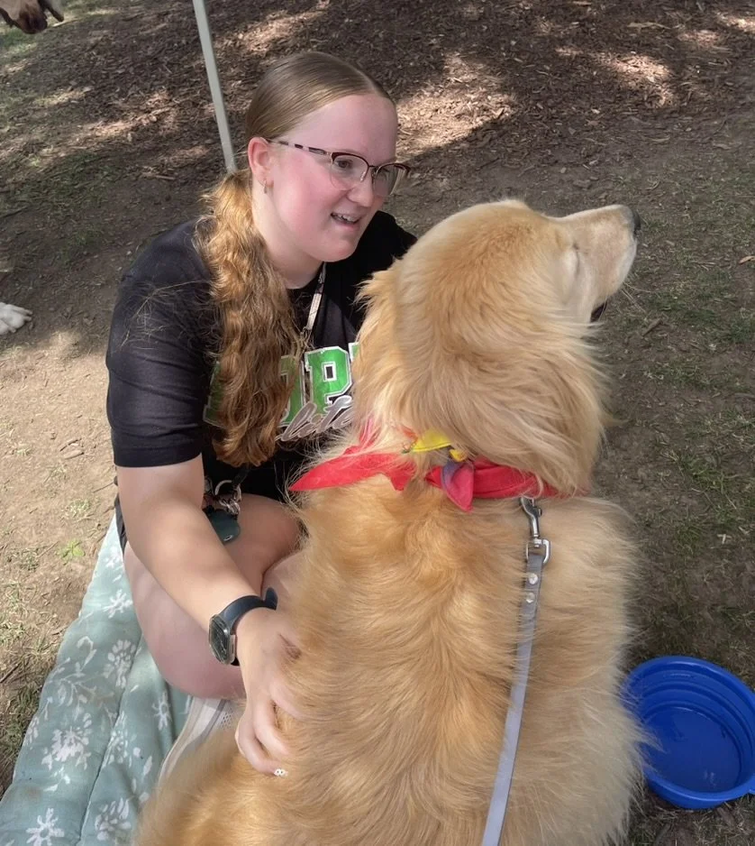 A young woman with glasses and long red hair kneels on the ground, smiling and petting a large golden retriever dog wearing a red bandana. They are outdoors, with the ground covered in dirt and some shaded areas.