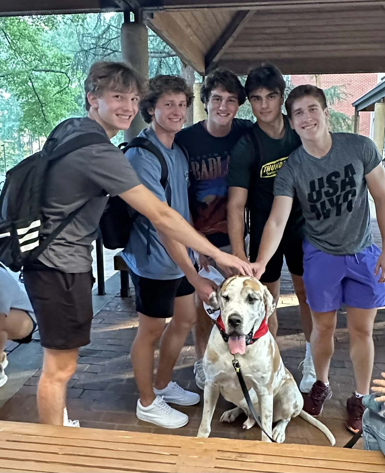 Group of five young men with a large white and tan dog, all smiling, on a wooden porch with trees in the background.