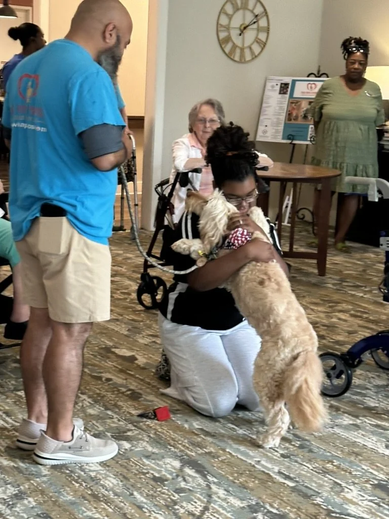 A woman kneeling on the floor hugging a large golden retriever puppy, with a man standing nearby and a woman in the background. The setting appears to be indoors, with a clock on the wall and a poster display behind.