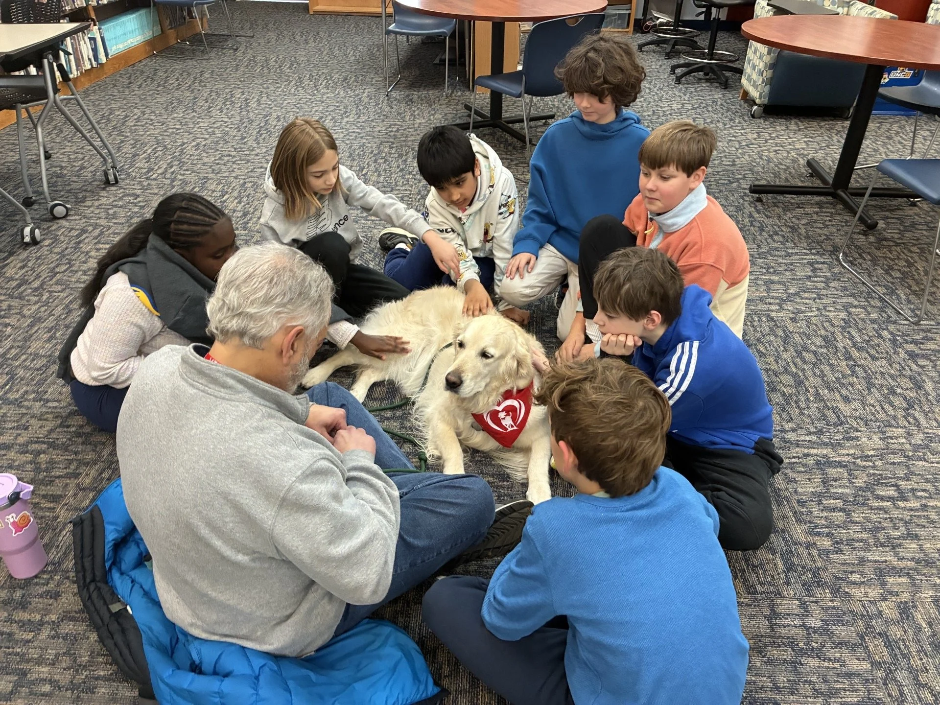A group of children and two adults gathered around a golden retriever dog lying on the floor in a library or classroom setting. The children are petting and looking at the dog, which is wearing a red scarf with a white heart.