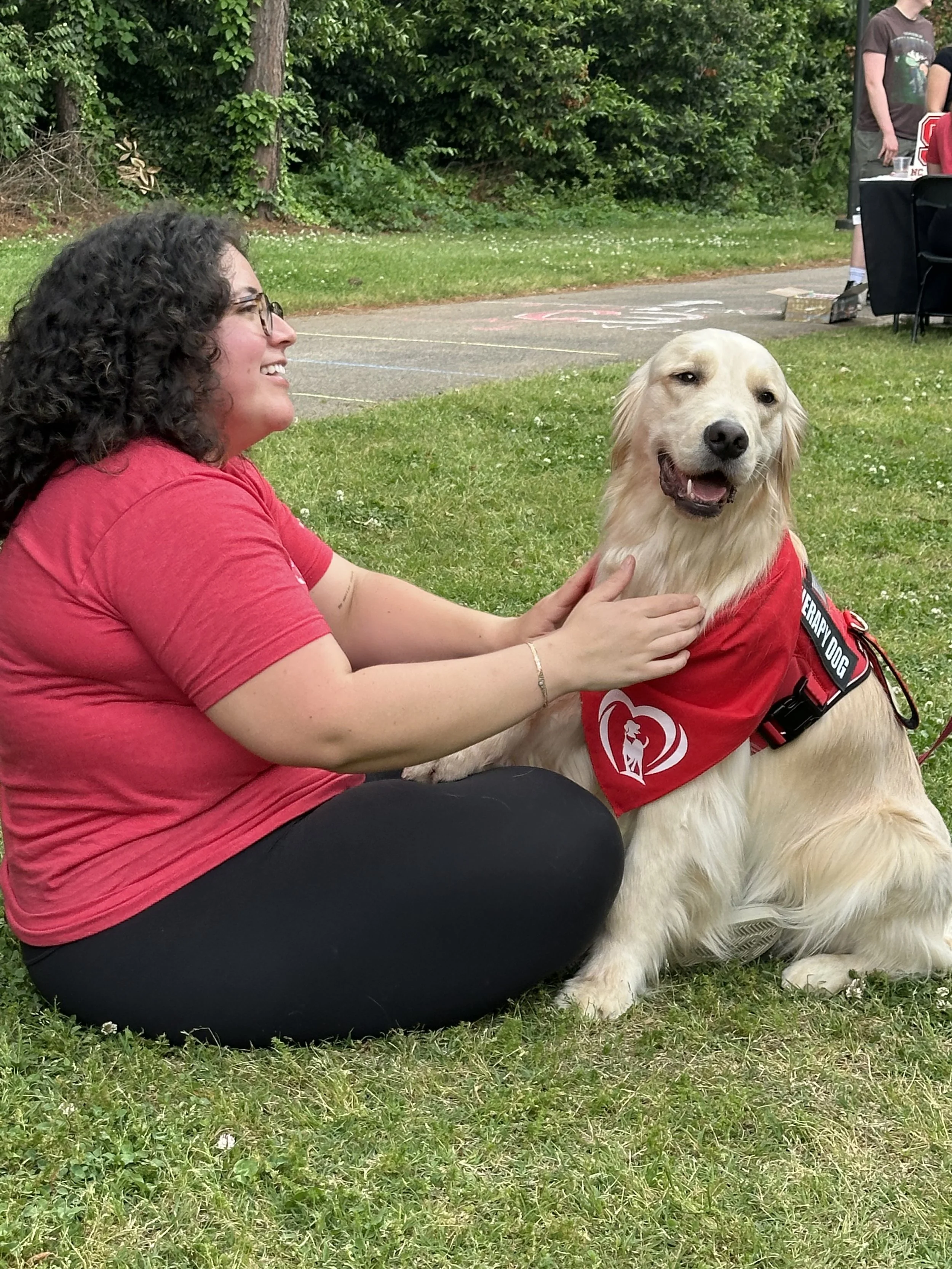 A woman sitting on the grass petting a golden retriever dog wearing a red vest with a therapy dog patch at an outdoor event.
