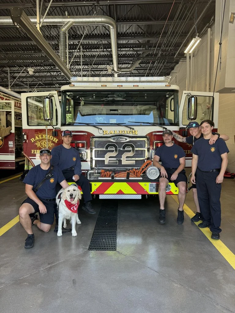 Group of four firefighters and a service dog posing in front of a fire truck inside a fire station garage.