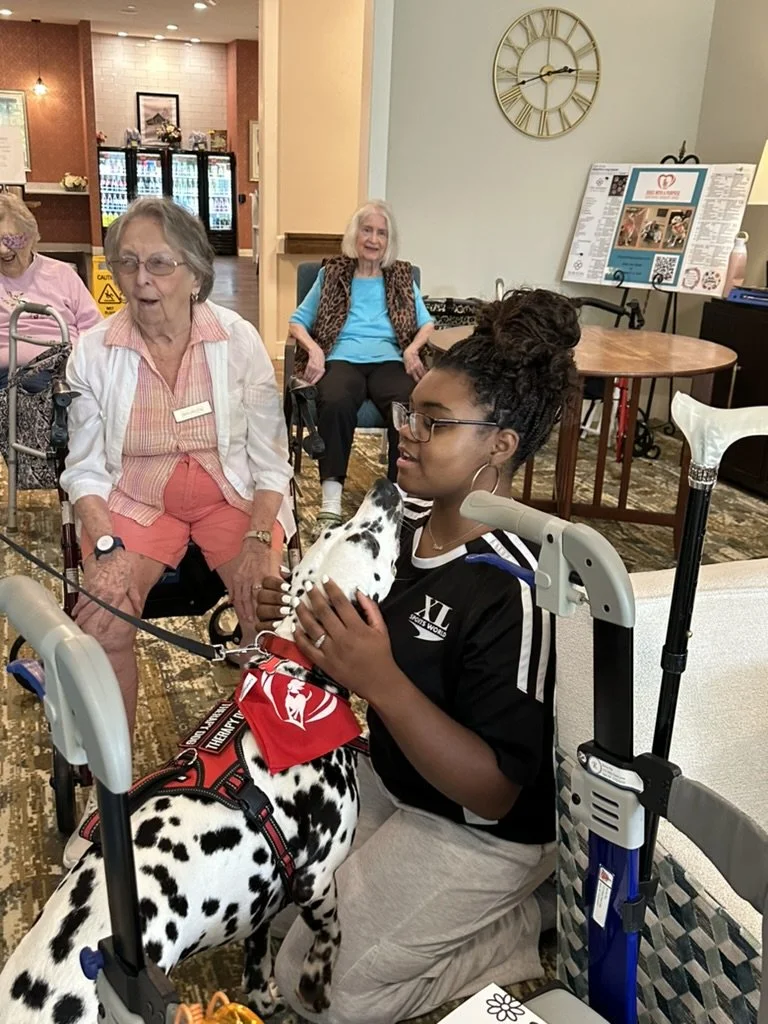 Young woman holding a Dalmatian puppy in an indoor facility with elderly women in wheelchairs nearby.