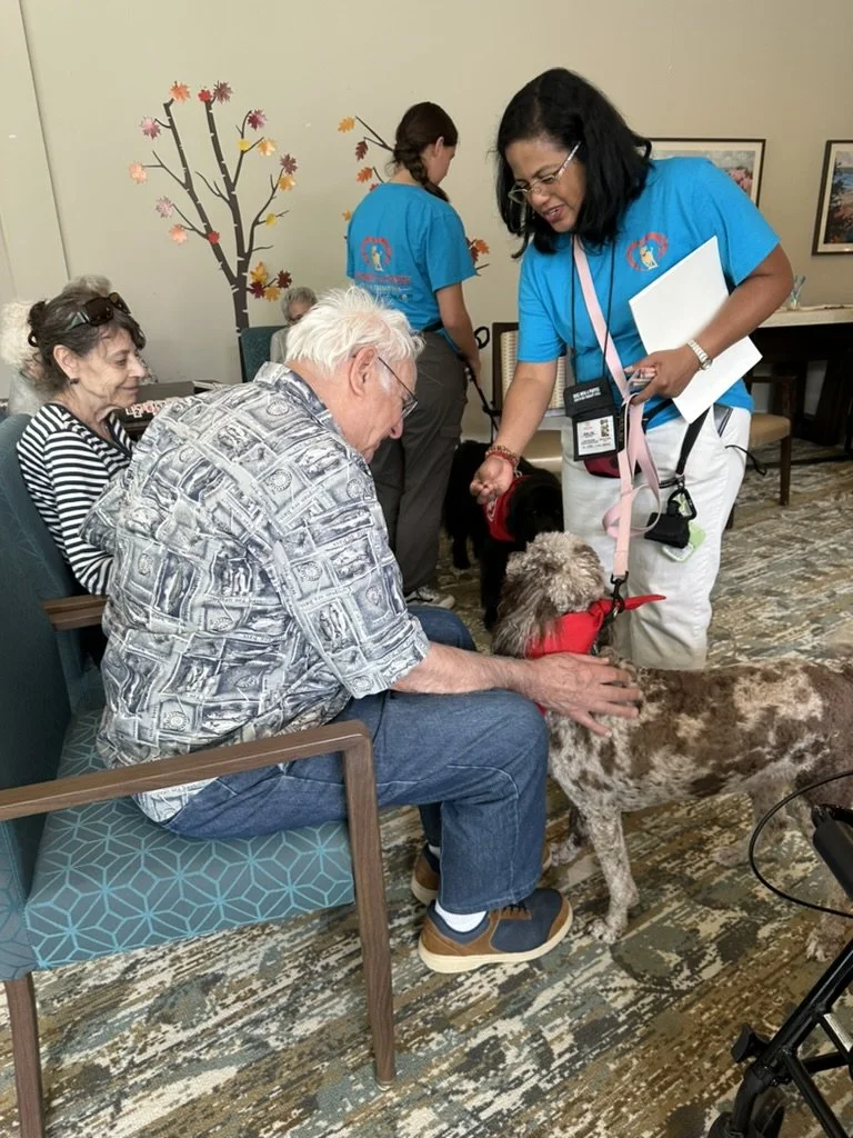 Older man with white hair and glasses sitting on a blue patterned chair, petting a brown and white dog with a red bandana, while a woman in a blue shirt interacts with the dog. Two women in blue shirts stand in the background with a wall decoration o