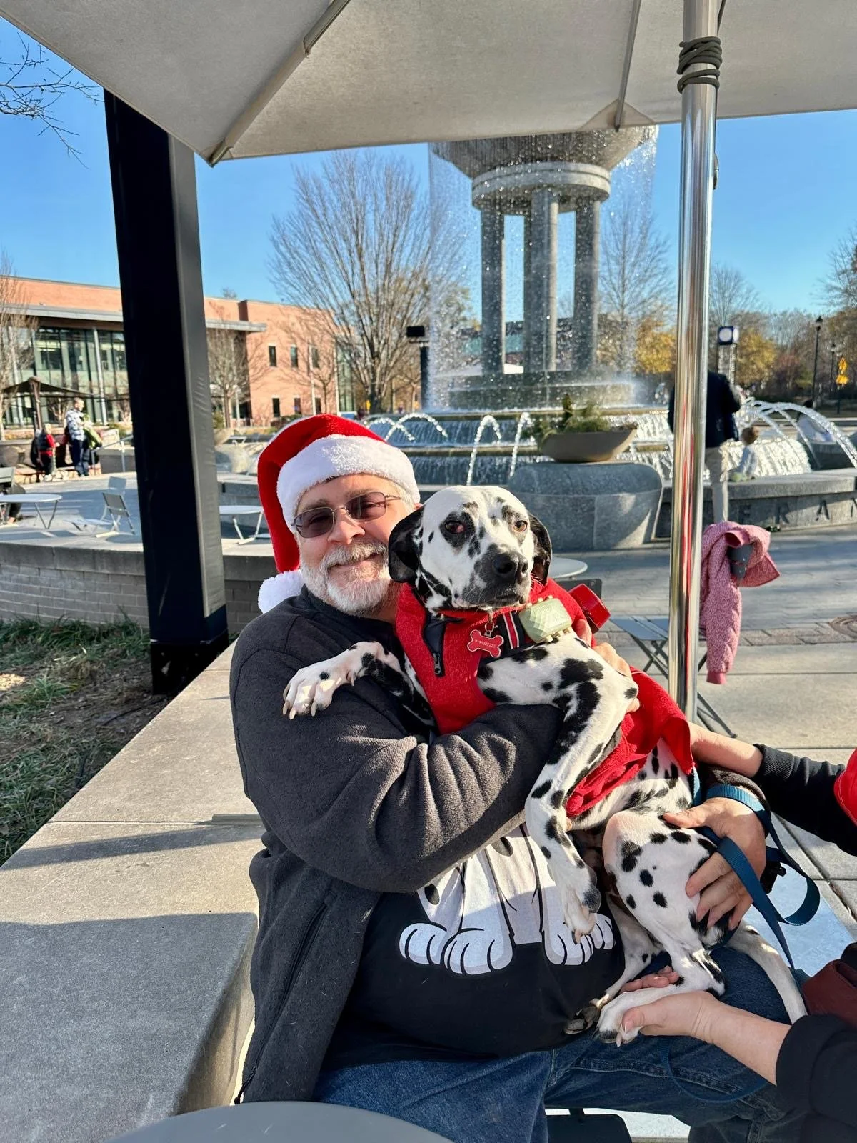 A man wearing a Santa hat is holding a Dalmatian dog dressed in a red jacket, sitting on his lap. They are outdoors near a fountain with a large water tower in the background, under a sunny blue sky.