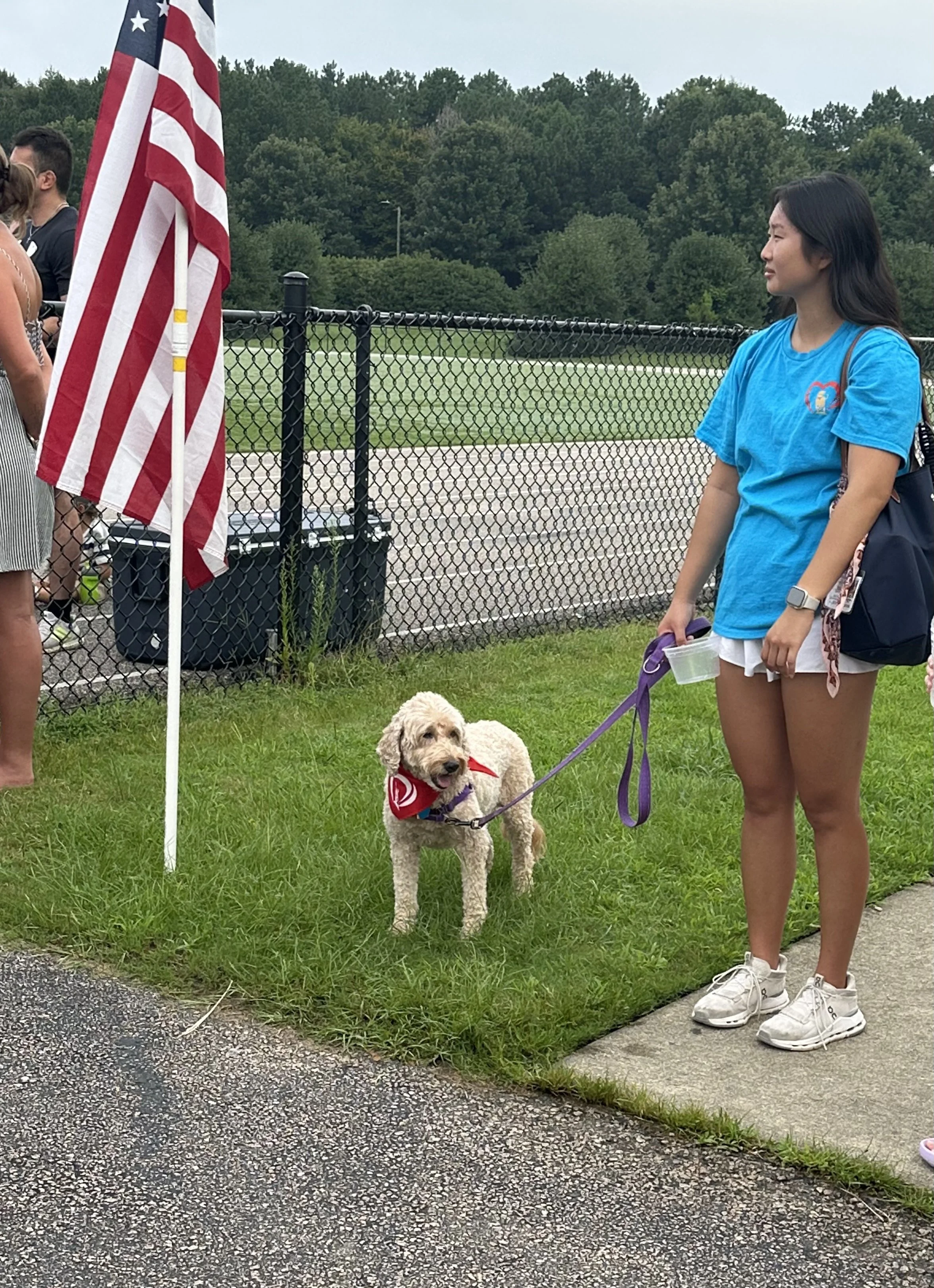 A young woman stands outdoors on a sidewalk, holding a purple leash attached to a small dog with curly beige fur, wearing a red bandana. The woman is dressed in a blue T-shirt, white shorts, and sneakers. She has a backpack and is holding a clear plastic cup. Behind her, there is a chain-link fence, an American flag, and a background of trees and cloudy sky.