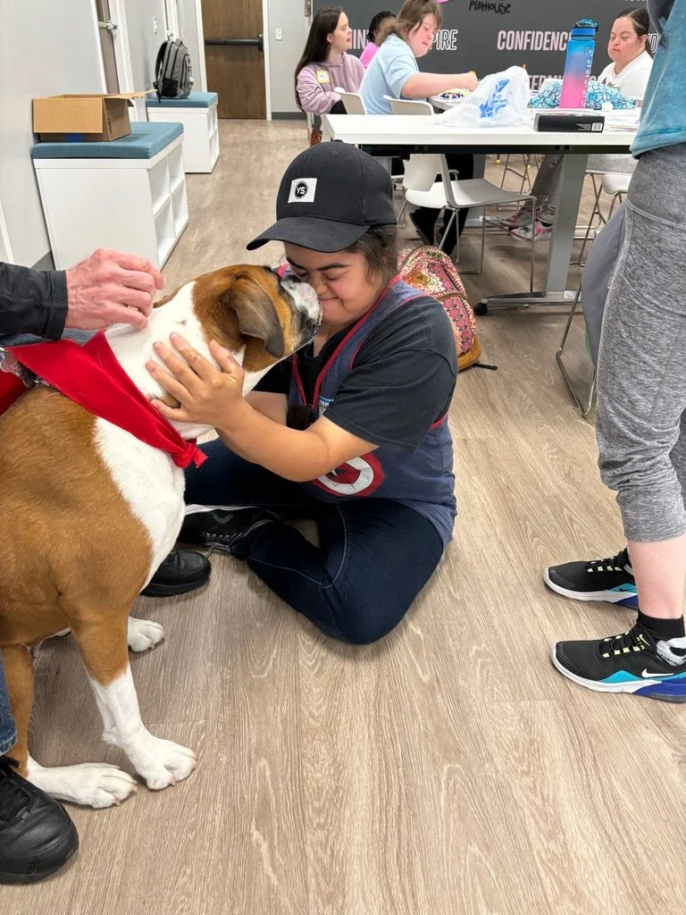 A young girl in a baseball cap and dark t-shirt squats on the floor hugging and kissing a large brown and white dog wearing a red bandana. The scene appears to be inside a classroom or community center with other children and tables in the background