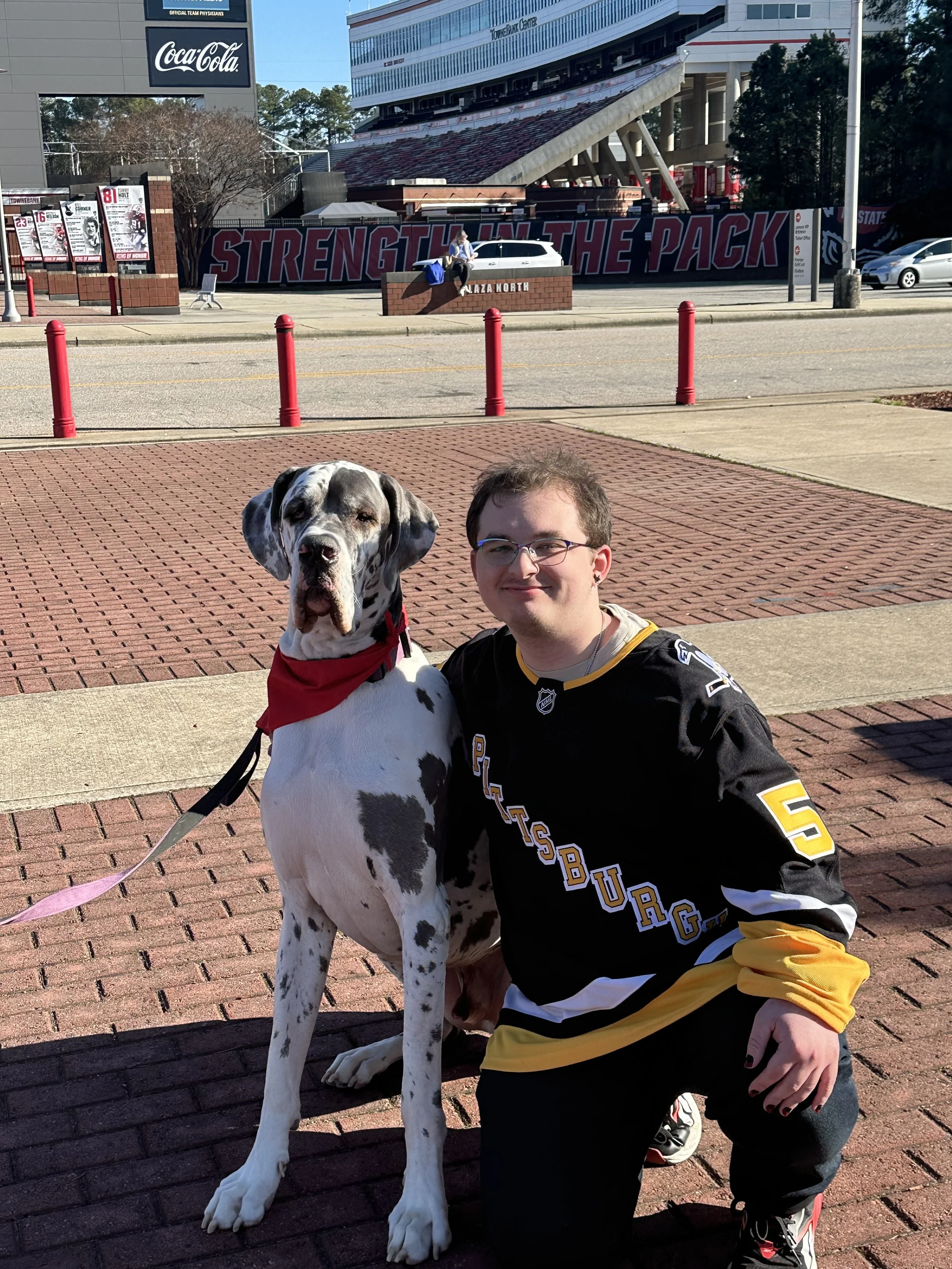 A man wearing a Pittsburgh Steelers jersey and glasses kneeling next to a Great Dane dog with a red bandana. They are outside in front of a building with a sign that says 'Strength in the Pack' and a brick fence with plaques.