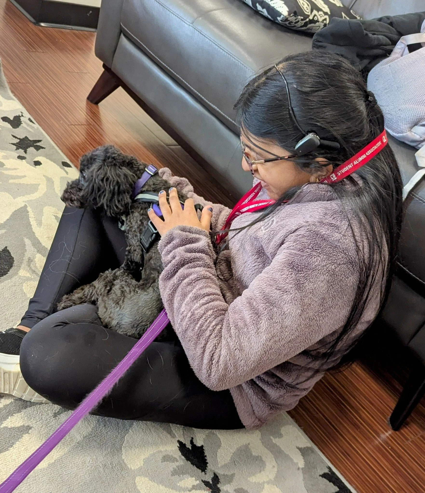 A woman with long dark hair and glasses lying on a couch, smiling while petting a black curly-haired dog with a purple collar. She is wearing a fuzzy brown jacket and a red lanyard around her neck, and there are black clothes and a patterned bag on t