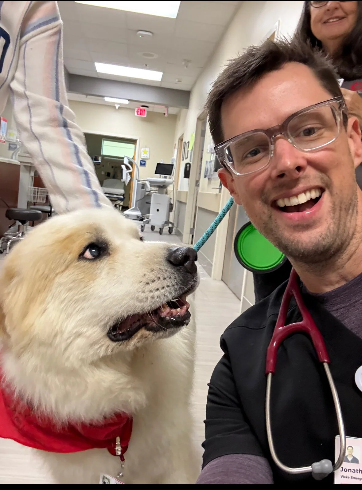A smiling man with a stethoscope around his neck taking a selfie with a golden retriever wearing a red bandana in a hospital or clinic corridor. An overmedicated woman is partially visible in the background.