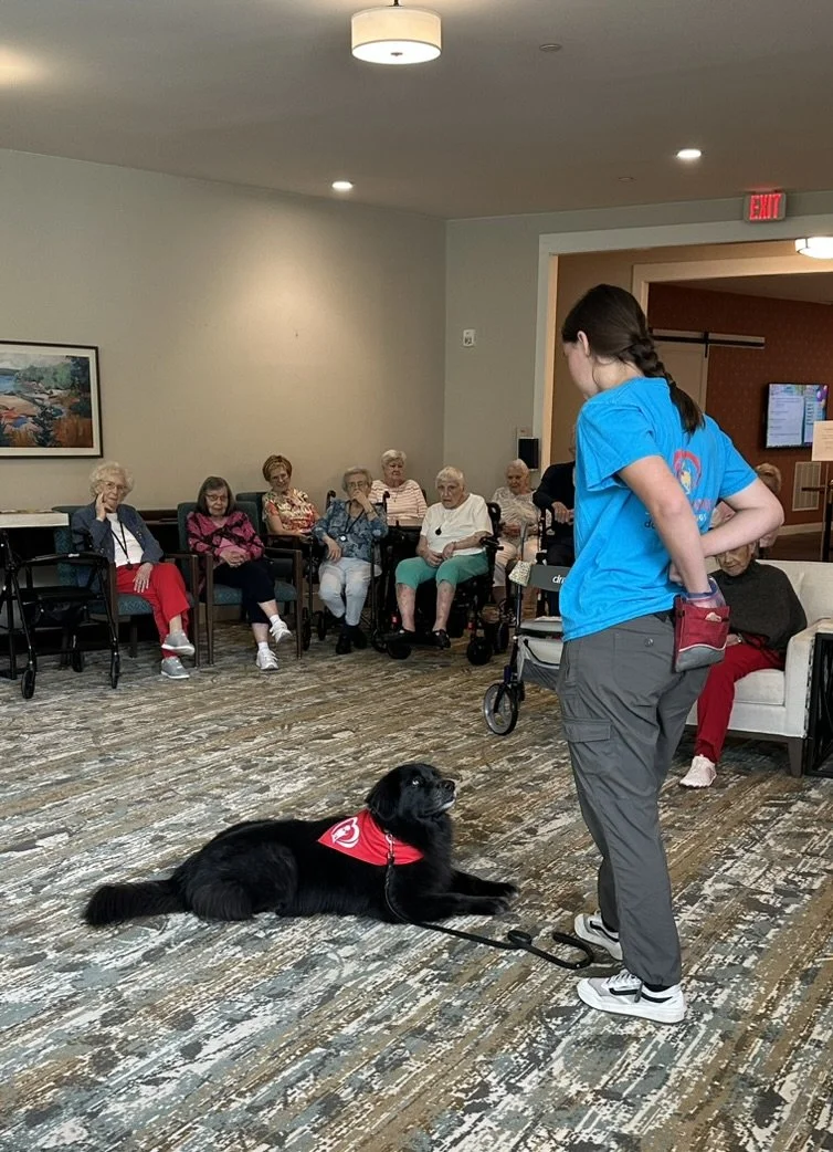 A young woman in a blue shirt and gray pants stands in front of an audience of elderly people in a community room. She has a black guide dog with a red vest lying on the floor at her feet. The elderly audience is seated, watching the woman and her do