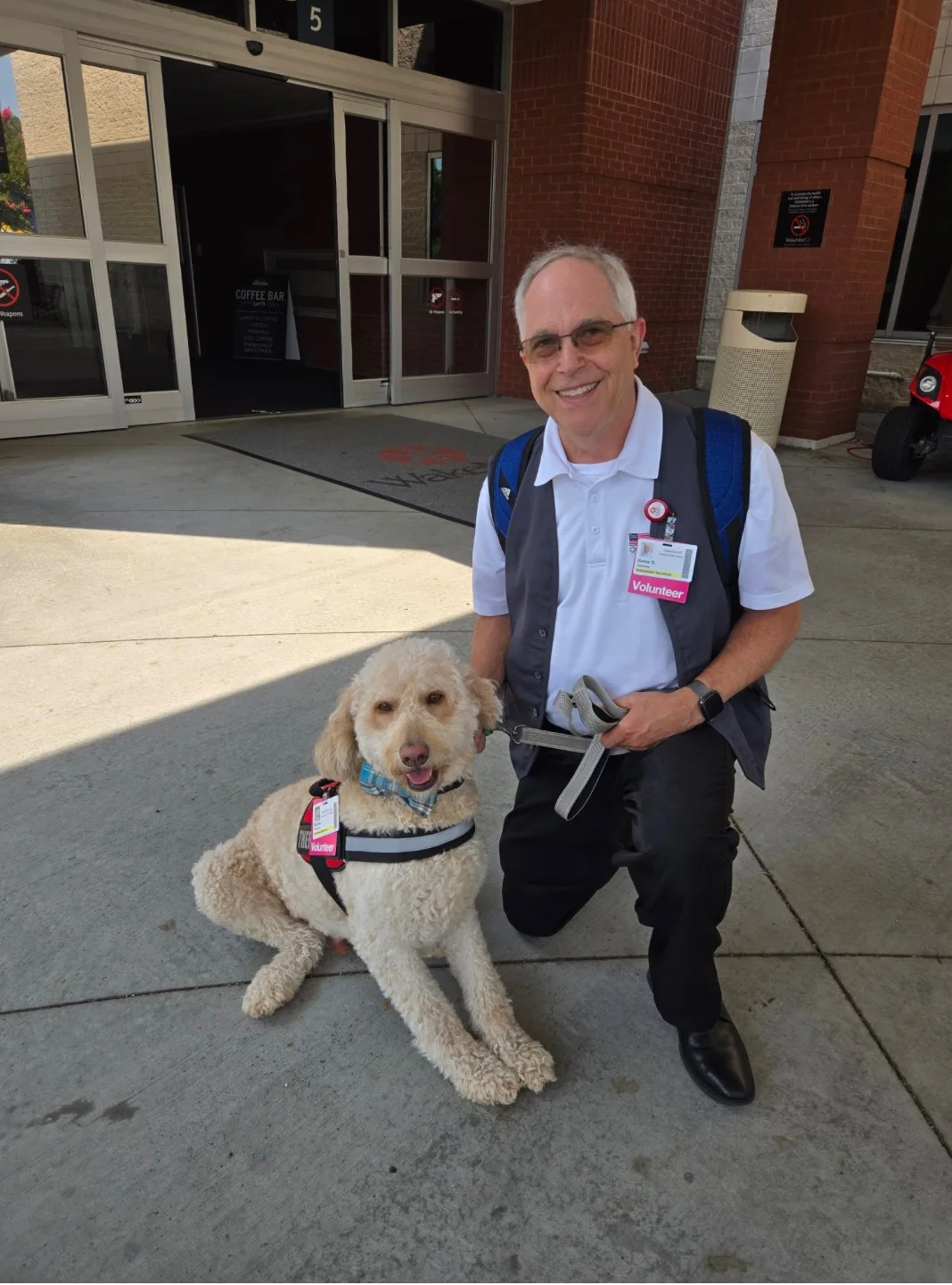 A smiling man in volunteer attire kneeling beside a service dog outside a building.