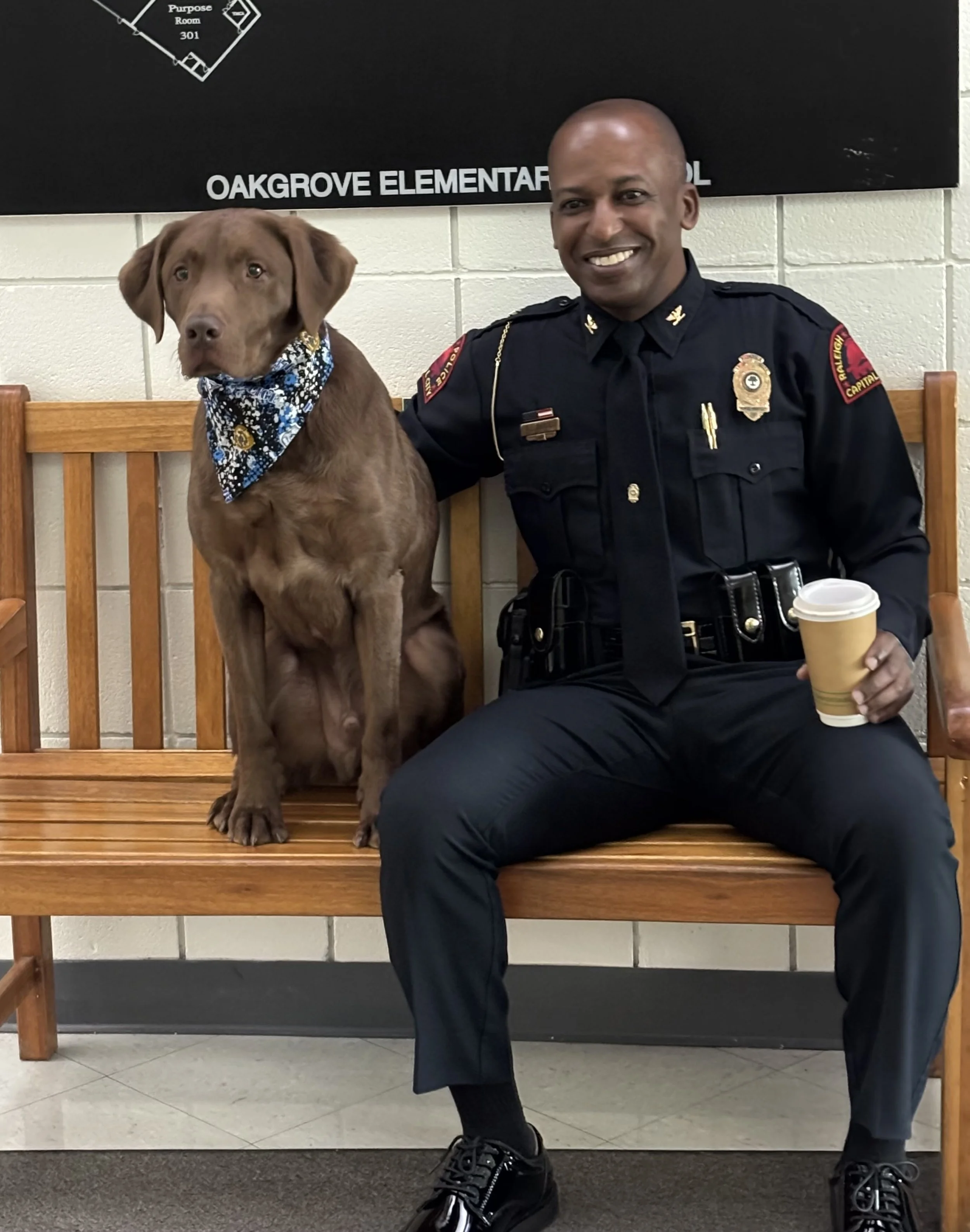 A man in a police uniform sitting on a wooden bench, holding a coffee cup in his right hand, with a brown dog wearing a bandana sitting on the same bench on his left. They are in a room with white tiled walls and a black sign that reads 'OAKGROVE ELEMENTARY'.