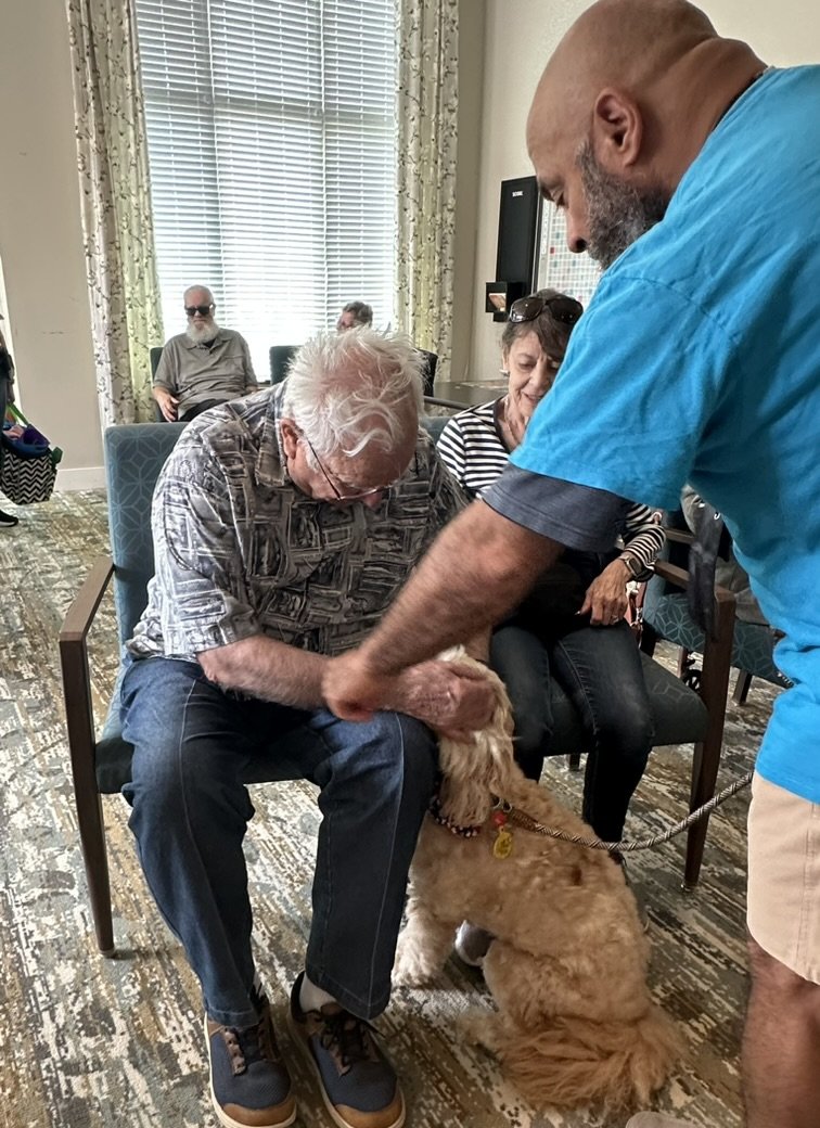 An elderly man with white hair and glasses sitting in a chair, petting a brown dog with a collar, while a man in a blue shirt and tan shorts holds the leash. Two women are sitting nearby, and a man with a beard and sunglasses is in the background, se