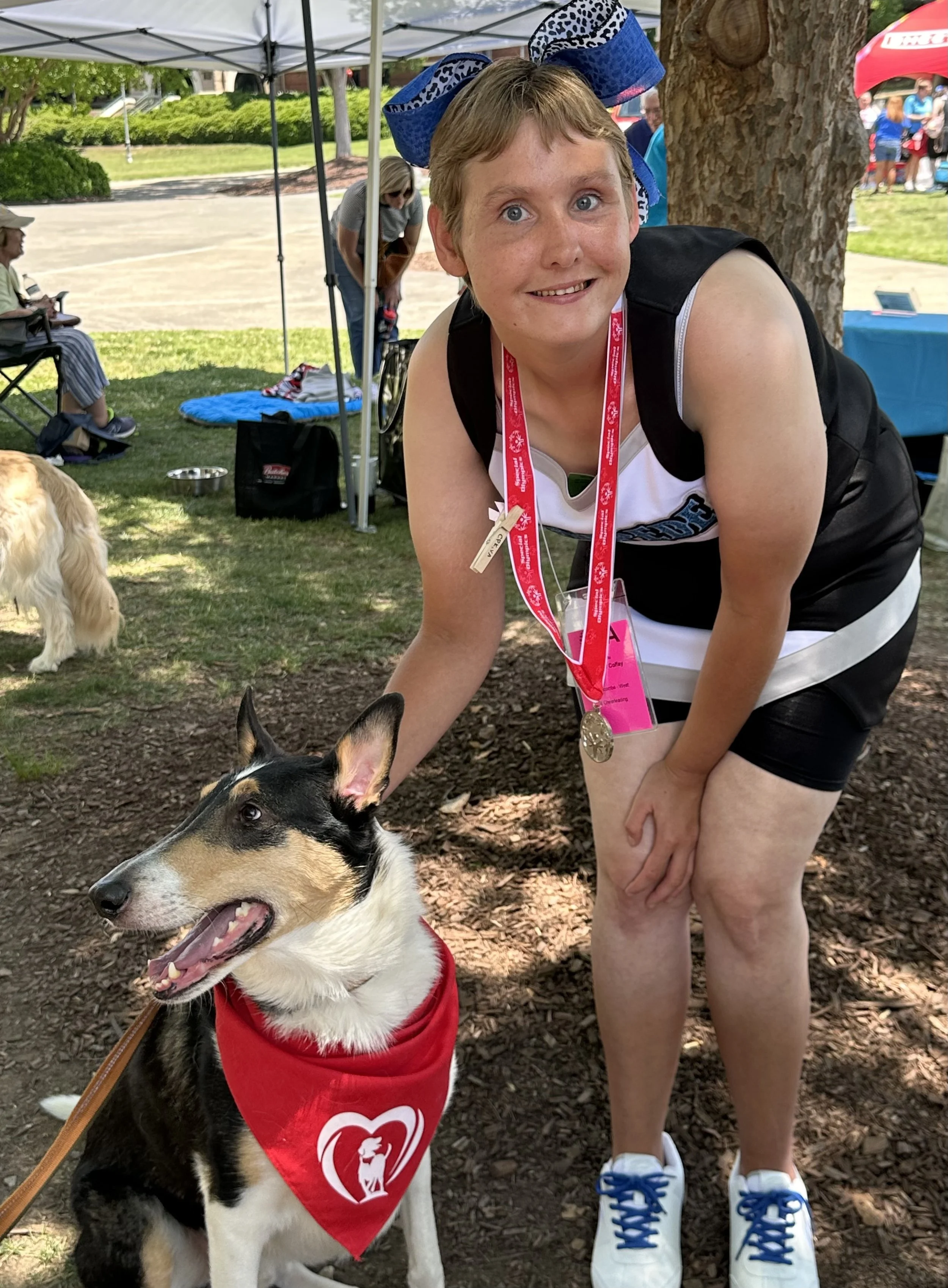 A woman wearing a black tank top and athletic shoes is smiling while posing with a black, white, and tan Border Collie dog wearing a red vest and a heart-shaped sign. The woman is also wearing a lanyard with a medal and an event card. There are peopl