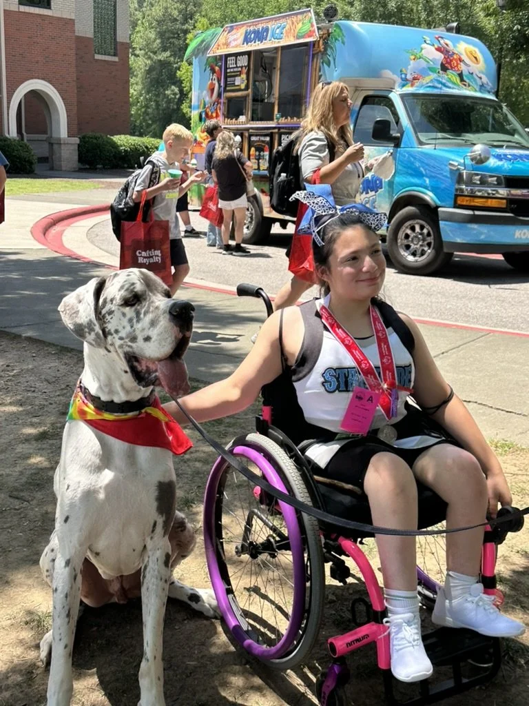 A young girl in a wheelchair with a spotted Great Dane dog sitting beside her outdoors at a sunny event. The girl is smiling, wearing a 'Stranger Things' shirt, a bow in her hair, and a pink lanyard. The dog is wearing a rainbow bandana, and people a