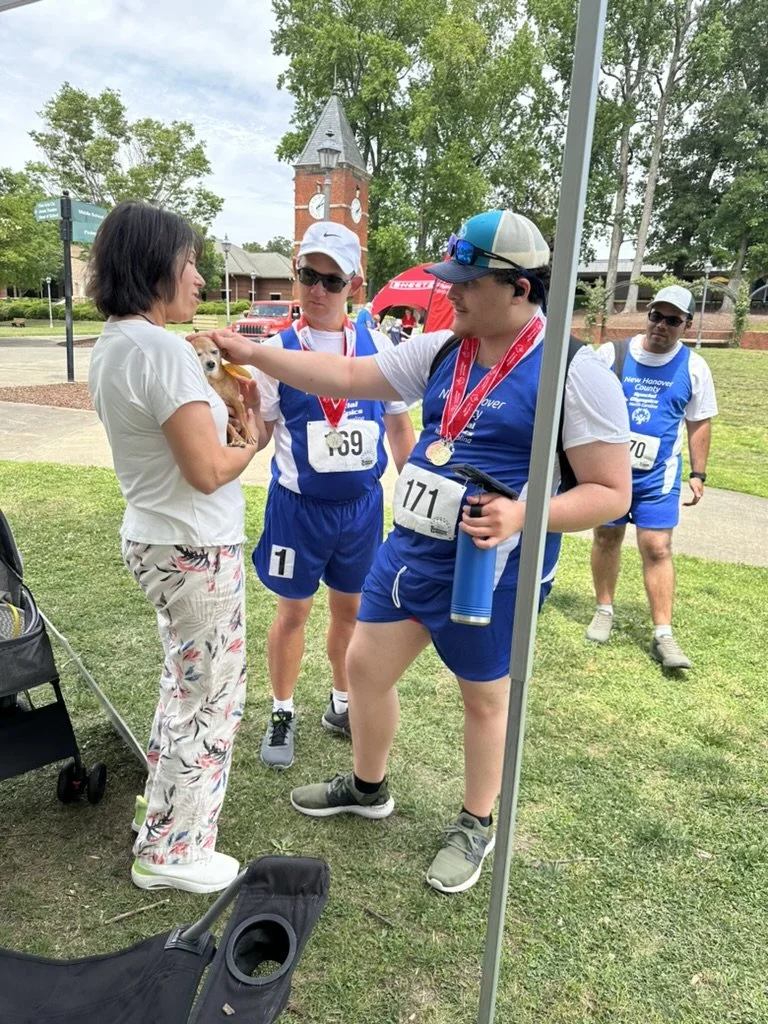 Four people at a race event, three men wearing blue sports gear with medals, one woman with dogs, and a clock tower in the background.