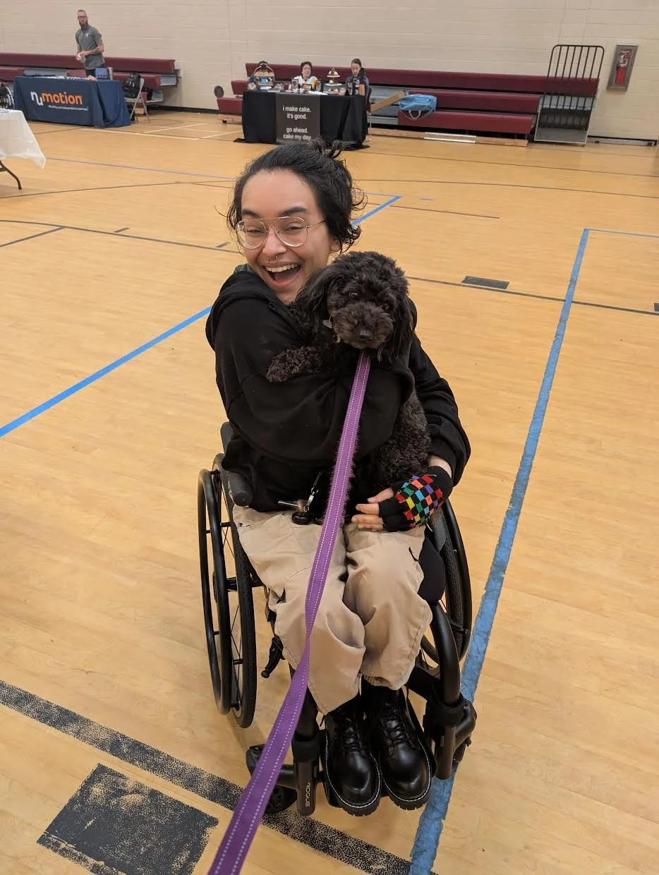 A woman in a wheelchair holding a black puppy with curly fur in an indoor gymnasium. She is smiling and wearing glasses, a black hoodie, and tan pants.