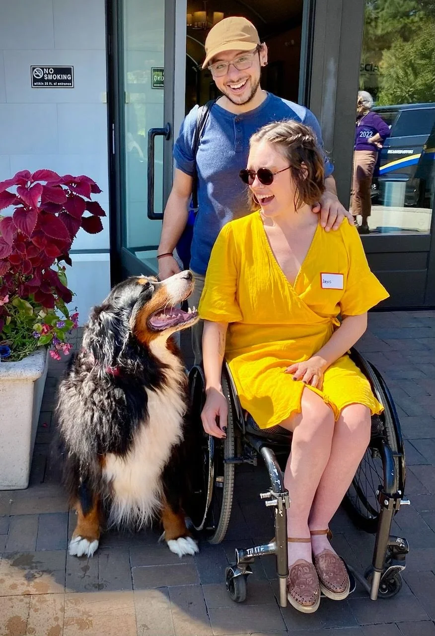 A woman in a yellow dress sitting in a wheelchair and smiling at a large happy Bernese Mountain Dog. A man with glasses, a hat, and a blue shirt stands behind her, also smiling. They are outside near the entrance of a building with flowers nearby.