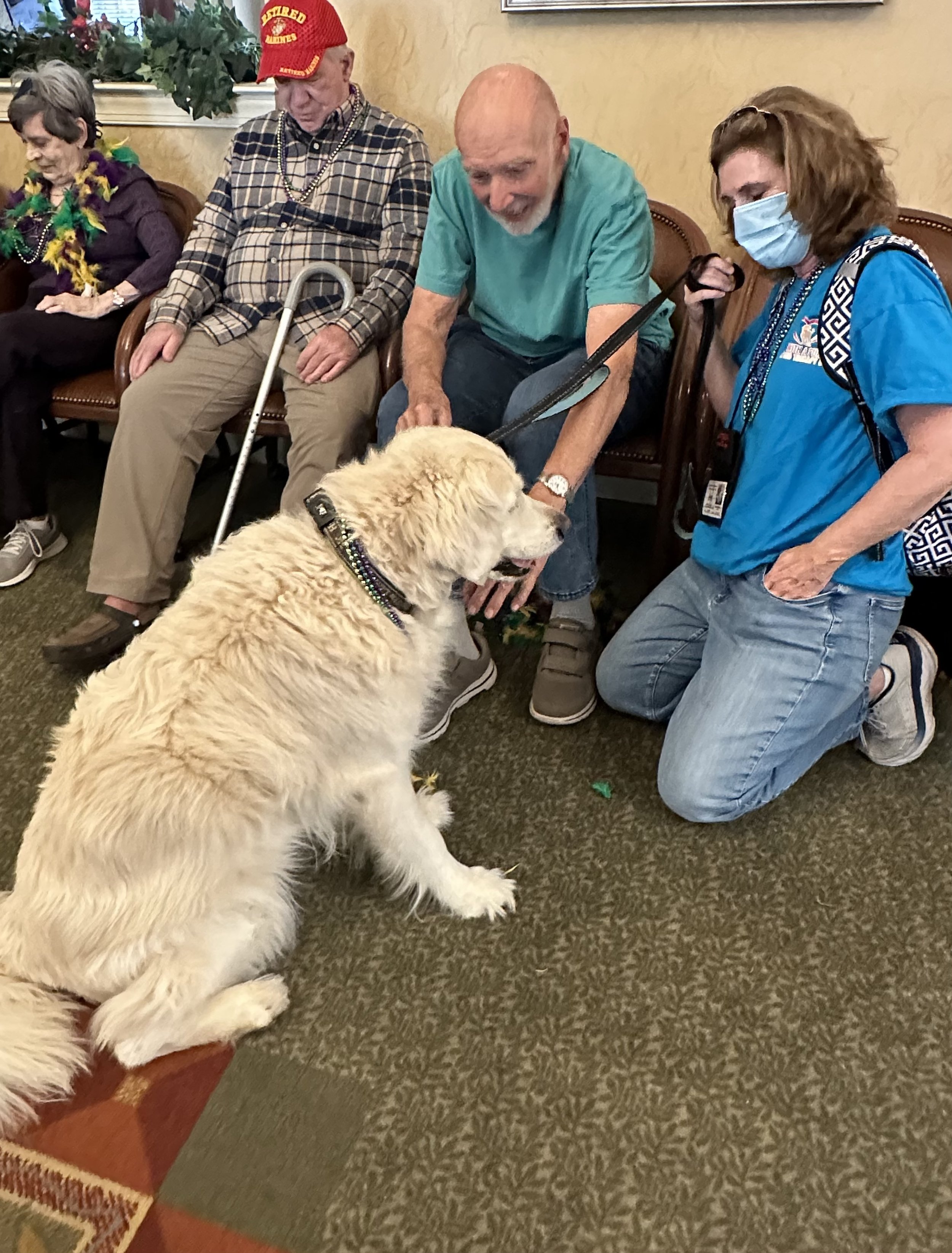 People sitting in a row, with the focus on an elderly man with a cane, a middle-aged man petting a large cream-colored dog, and a woman wearing a face mask. The group appears to be in a waiting area or community room.