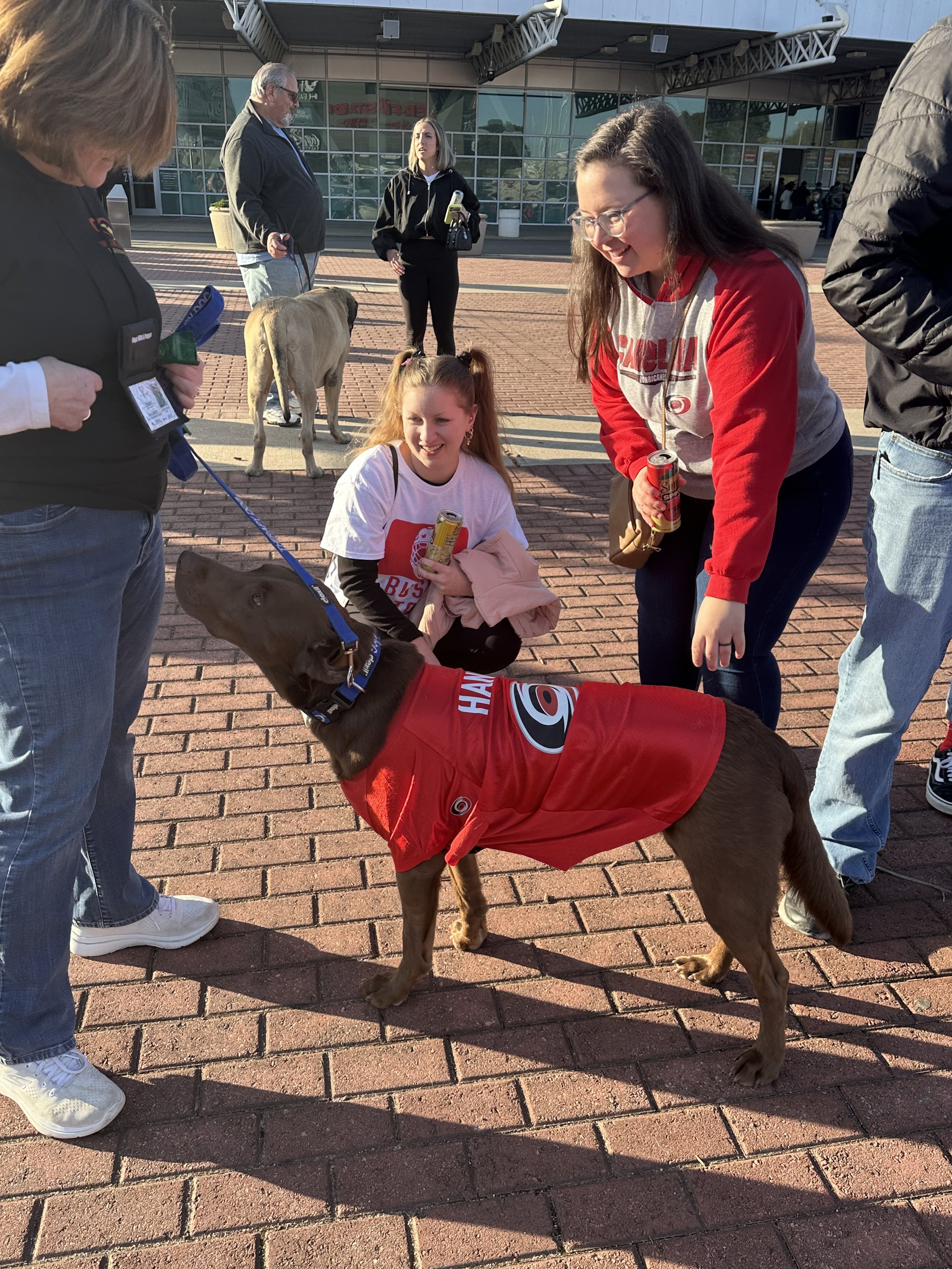 A brown dog wearing a red hockey jersey is on a leash, surrounded by people outside a building. Two girls are smiling at the dog, one with long brown hair wearing glasses and a gray and red shirt, and the other with red hair in pigtails, holding a ca