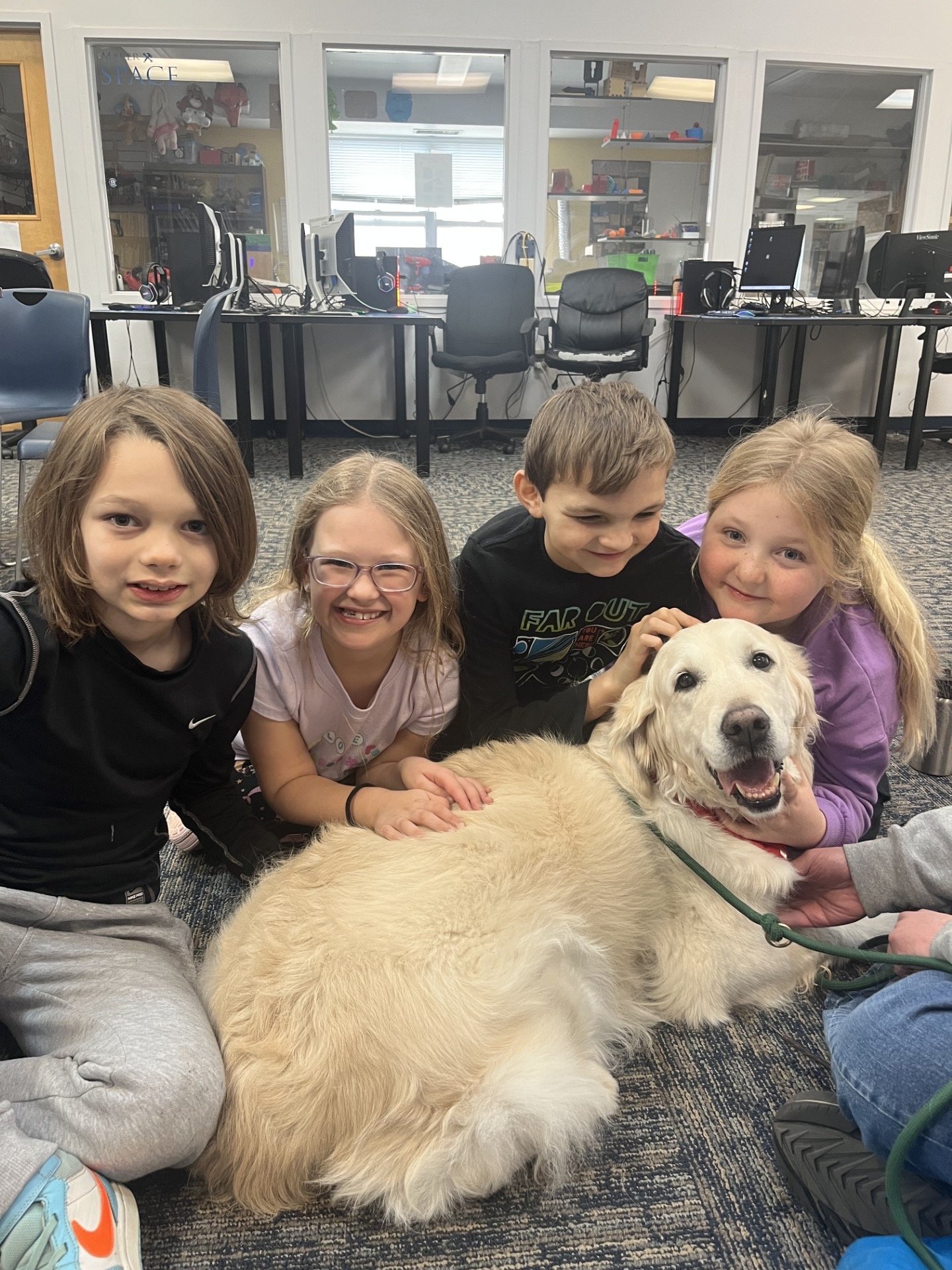 Four children and a dog sitting on the floor in a classroom, with computer desks and monitors in the background.