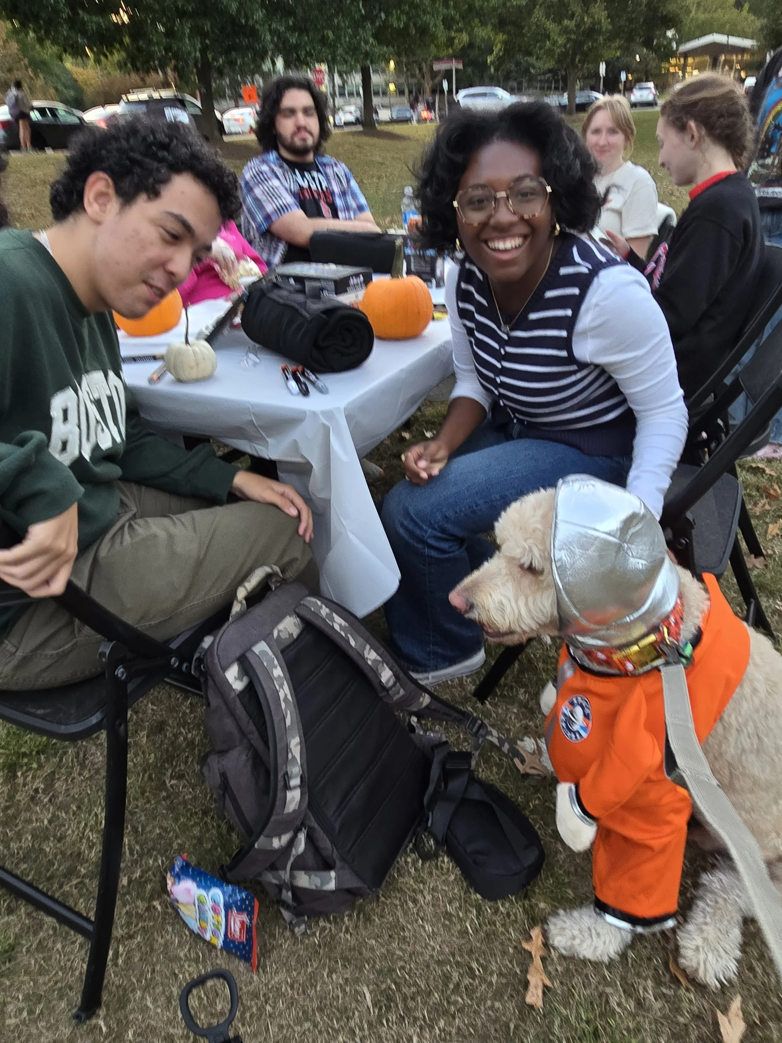 Group of people sitting at an outdoor table with pumpkins, backpacks, and snacks, with a service dog wearing an orange vest in the foreground.