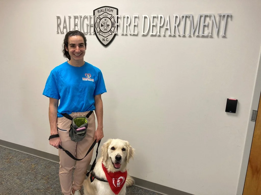 Smiling woman in scrubs standing with a service dog in front of a sign that reads 'Raleigh Fire Department'