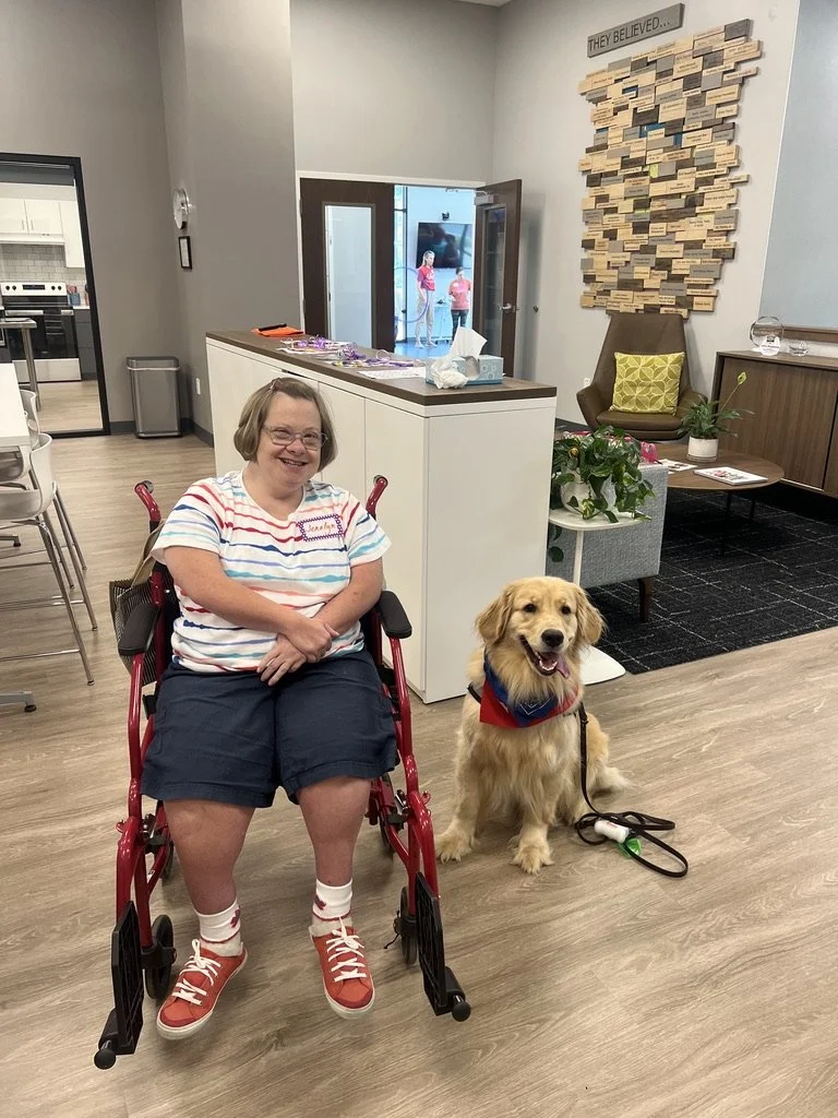 A smiling woman in a wheelchair with a service dog sitting beside her, both inside a room with light-colored walls and wooden flooring.