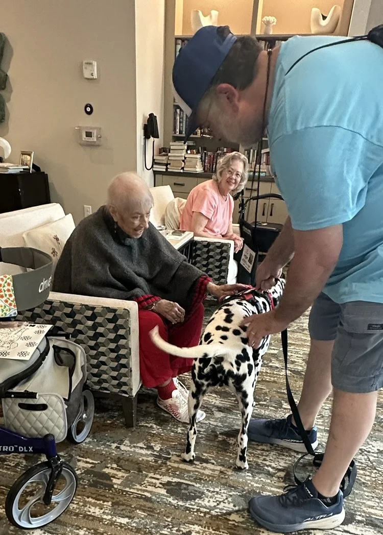 A person in a blue shirt and cap holding a Dalmatian dog on a leash in front of two elderly women sitting in a room.