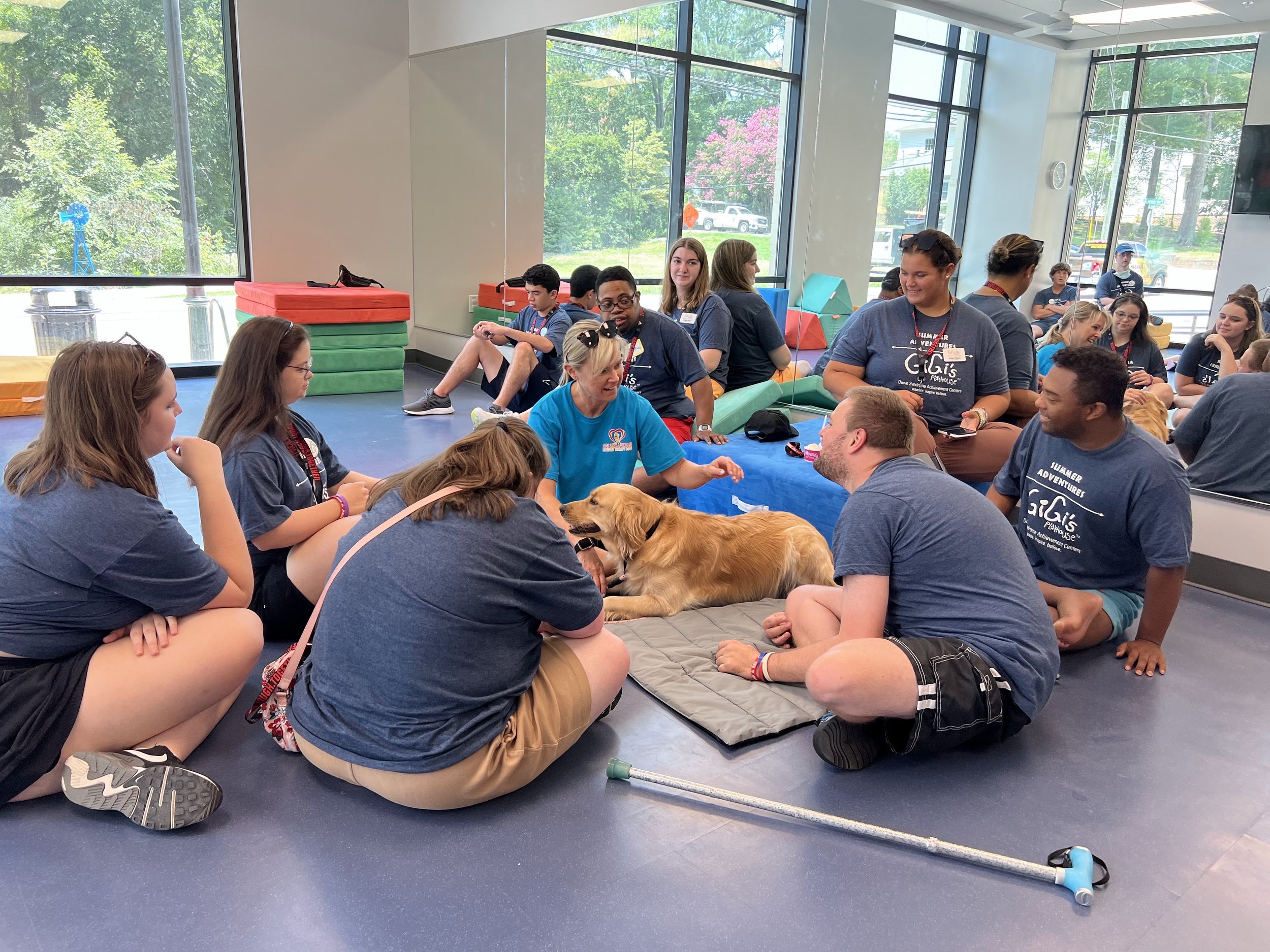 Group of young people sitting on the floor inside a room with large windows, engaging with a therapy dog and interacting with each other.