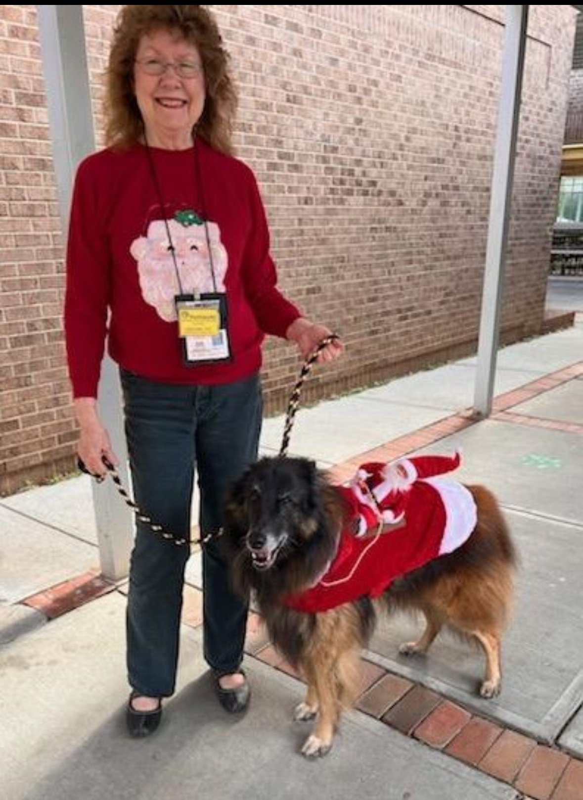 A cheerful woman wearing glasses and a red Christmas sweater with a Santa face, standing outdoors on a sidewalk, holding a dog on a leash. The dog is dressed in a red and white Christmas outfit, resembling Santa's costume.