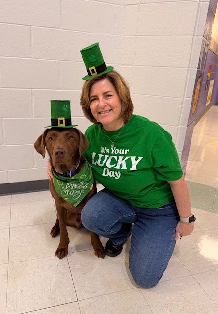 A woman dressed in a green St. Patrick's Day shirt and hat, smiling while kneeling next to a brown dog wearing a green bandana and a small green leprechaun hat.