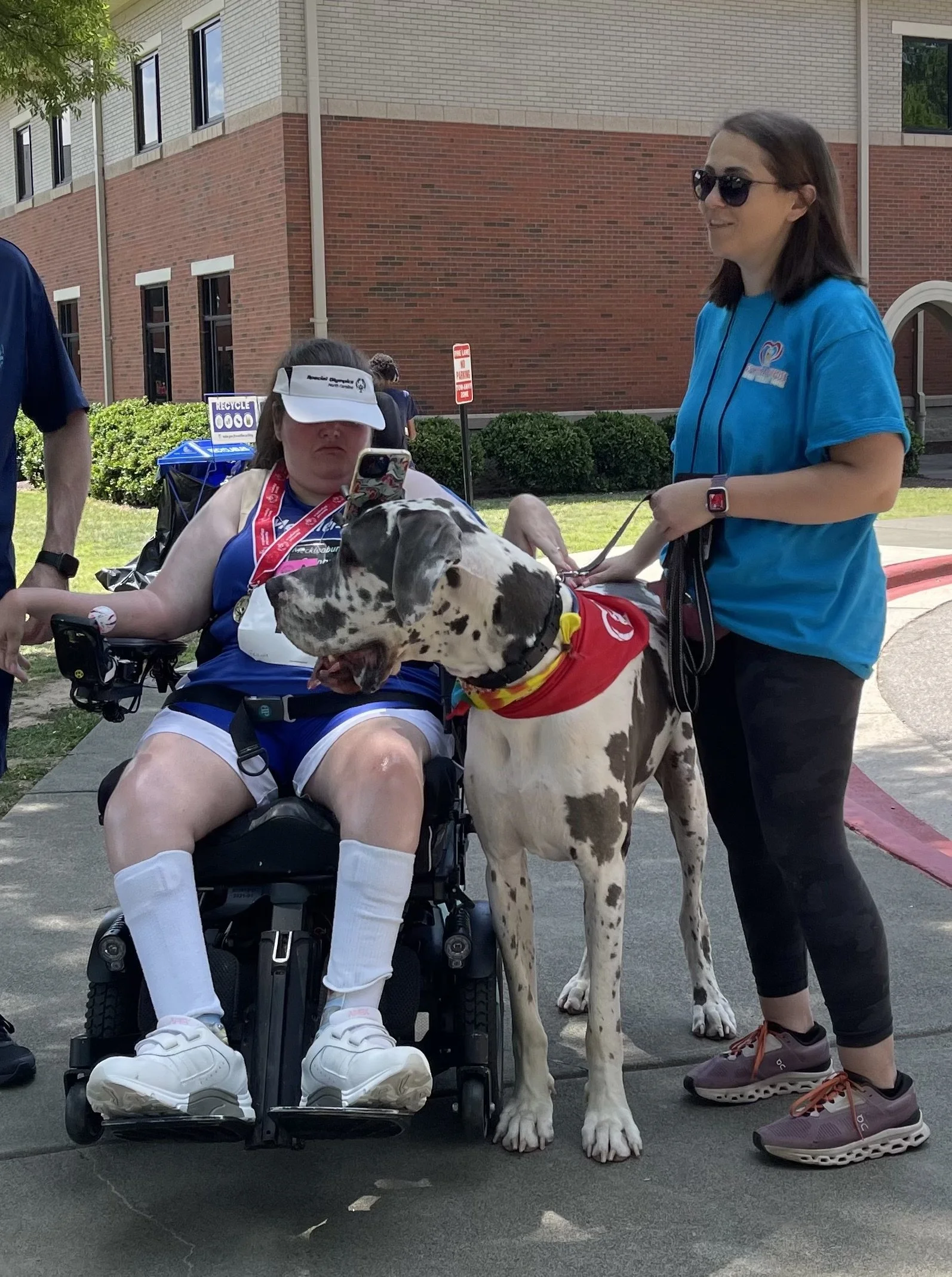A woman in a wheelchair, wearing a white visor and blue sportswear, is sitting outdoors with a large Dalmatian dog standing next to her. A woman in sunglasses and a blue shirt is standing nearby, holding the dog's leash. The background shows a brick 