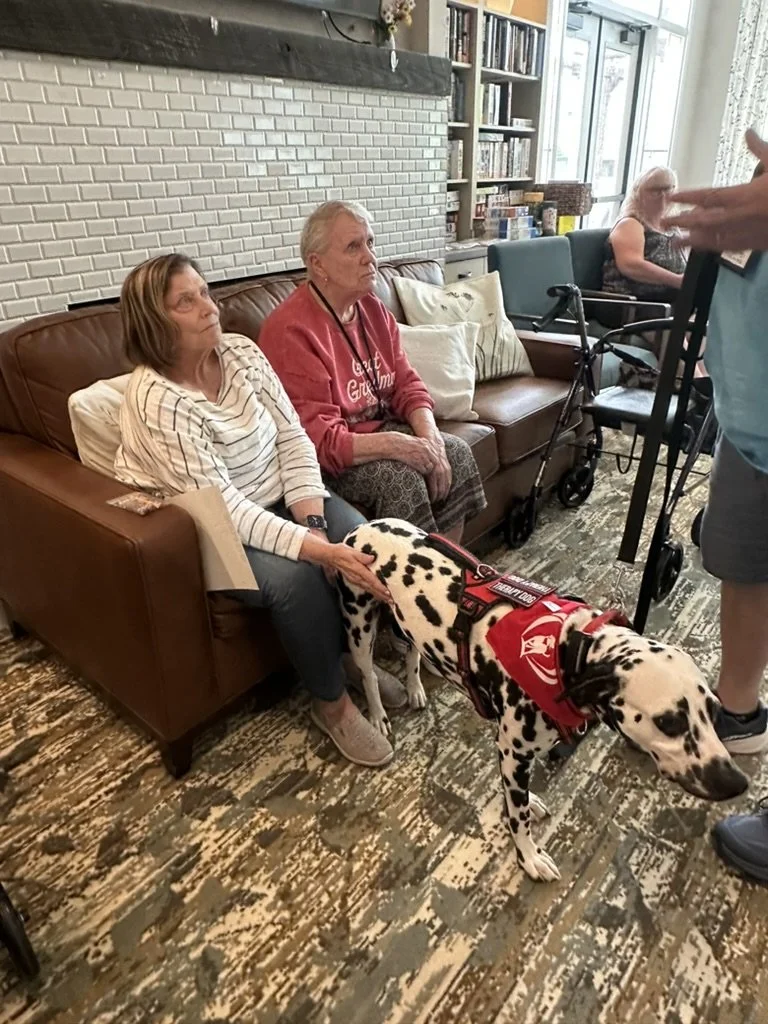 Two elderly women sitting on a brown couch in a library or community room, with a Dalmatian service dog in a red vest in front of them, and a person standing nearby.