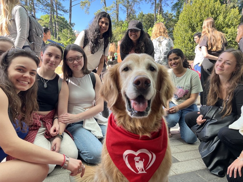 A group of women and young girls posing outdoors with a golden retriever dog wearing a red bandana with a logo. The group is sitting and standing on a paved area with trees and blue sky in the background.