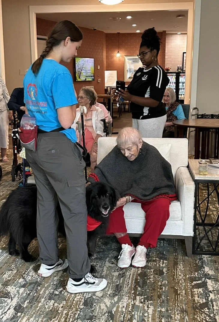 An elderly woman with grey hair sitting on a white armchair, smiling and petting a black service dog. A young caregiver in blue scrubs stands nearby, and a woman with glasses in a black shirt is using her phone in the background. The setting appears 