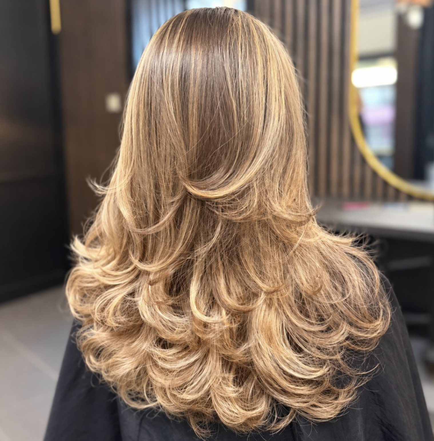 Back view of long, layered honey-blonde hair with voluminous curled ends in a salon