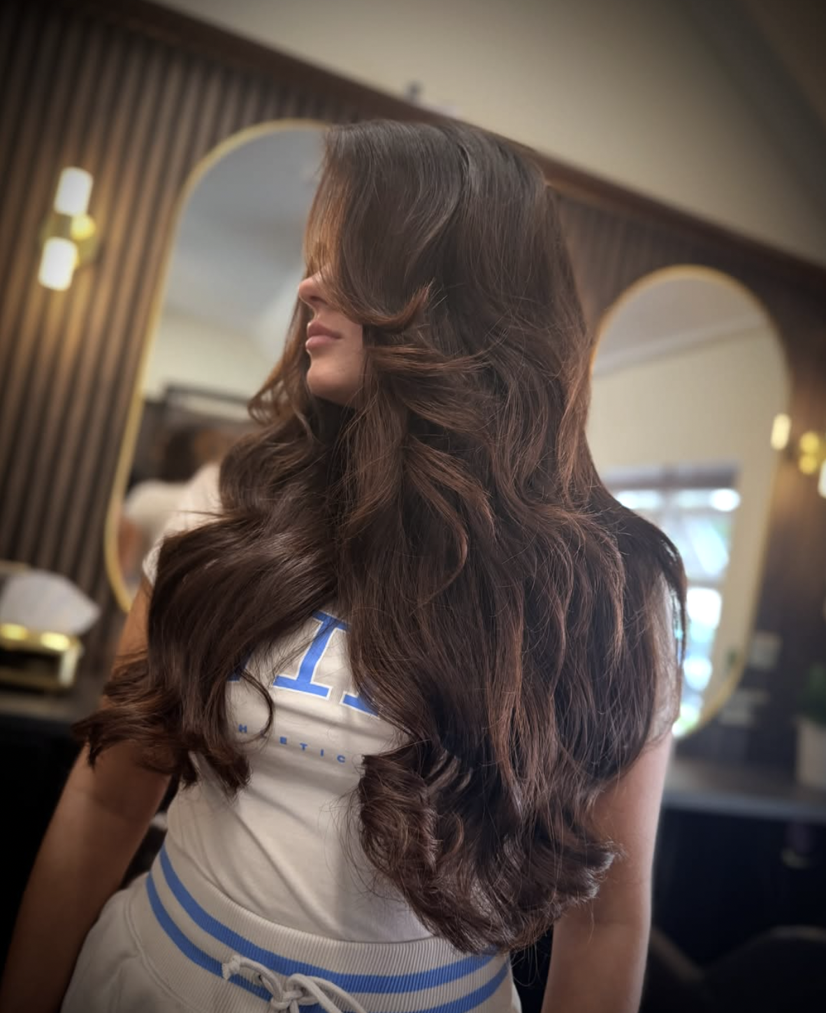 Woman with long, voluminous wavy brown hair posing indoors in front of arched mirrors