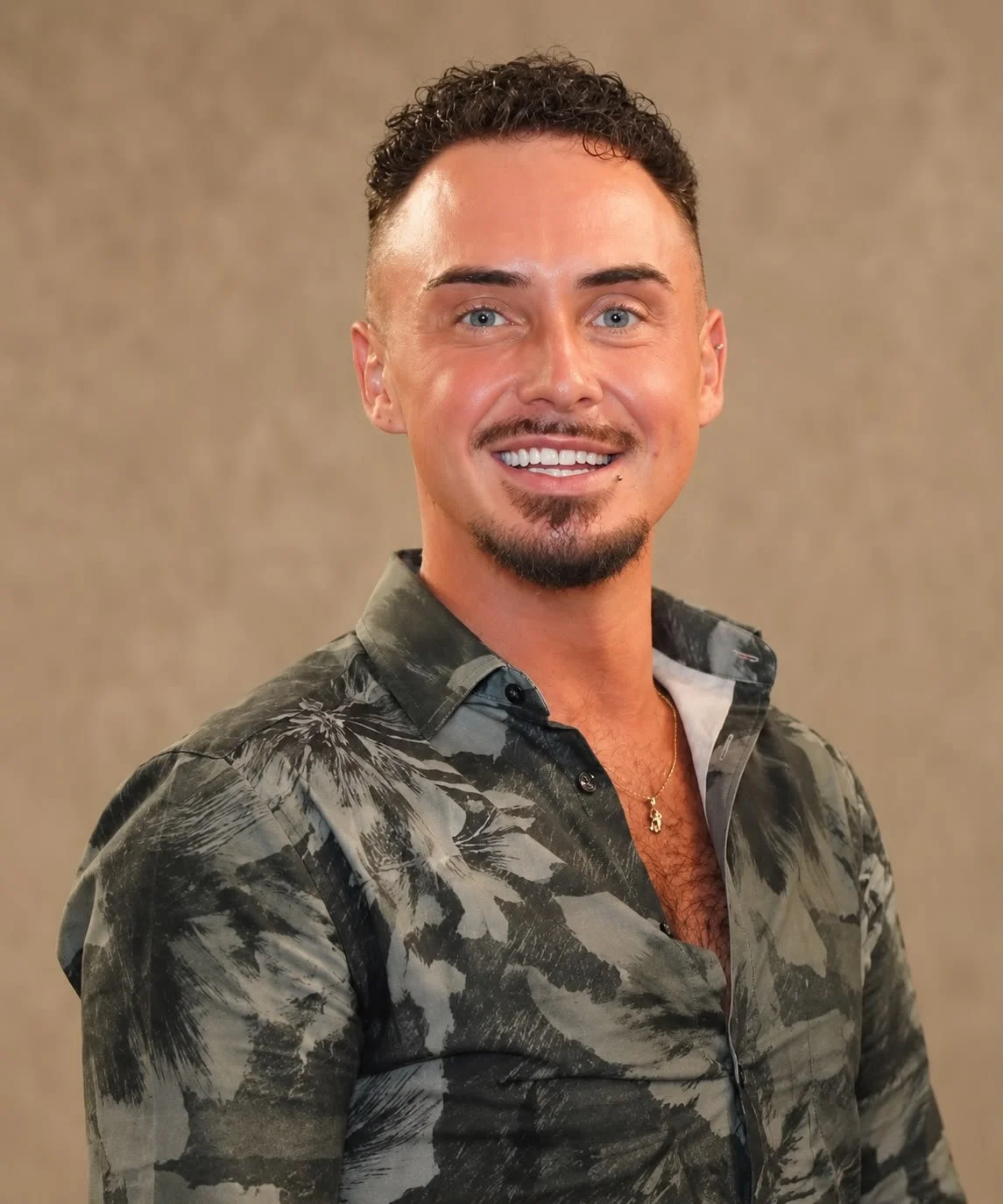 Smiling man with curly hair and goatee wearing a patterned button-down shirt against a beige background