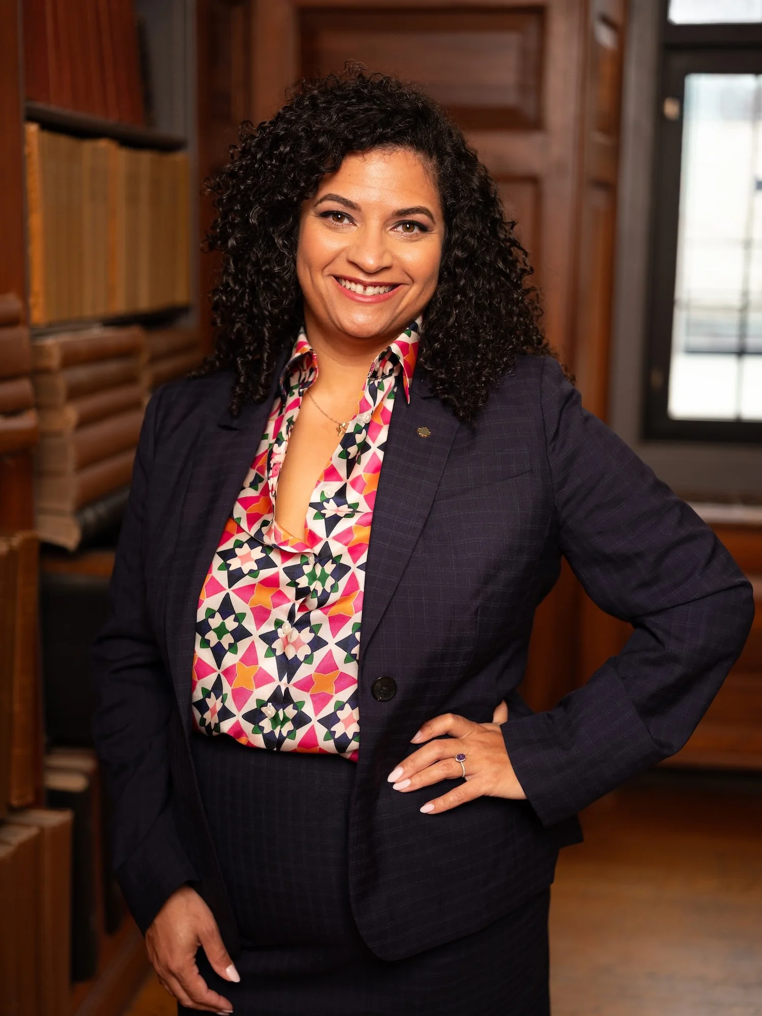 A woman with curly dark hair, smiling, wearing a colorful patterned blouse and a dark blazer, standing in a room with wooden bookshelves and a door with a window.