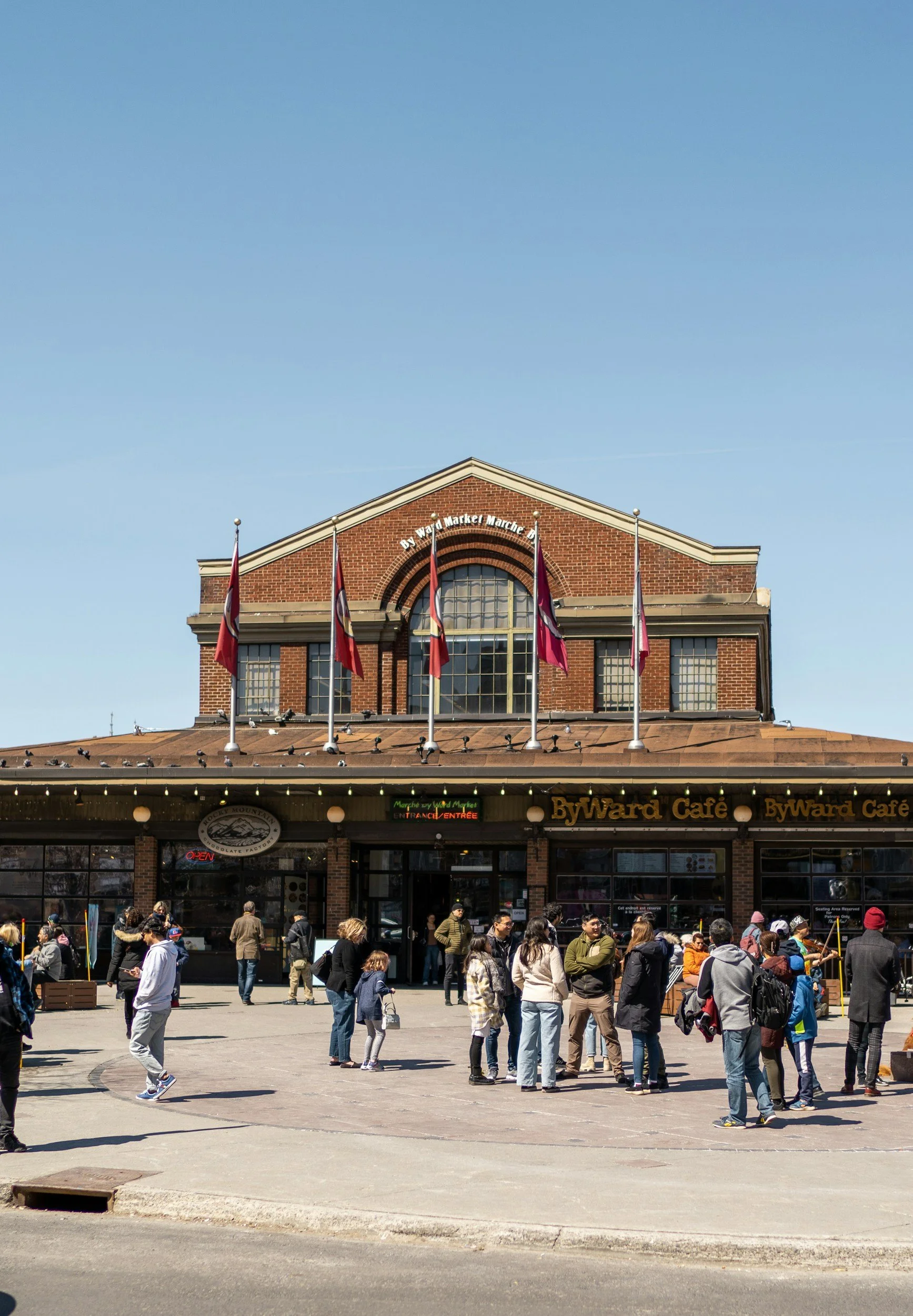 A busy outdoor scene in front of a brick building with a sign for Byward Market and a café, with people walking and standing around.
