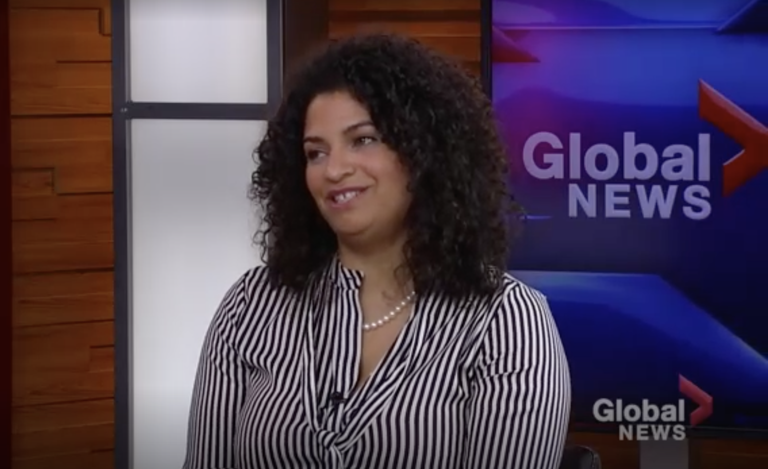 A woman with curly dark hair wearing a striped blouse and pearl necklace is on a TV news set with a background that includes a purple and red logo that says Global News.