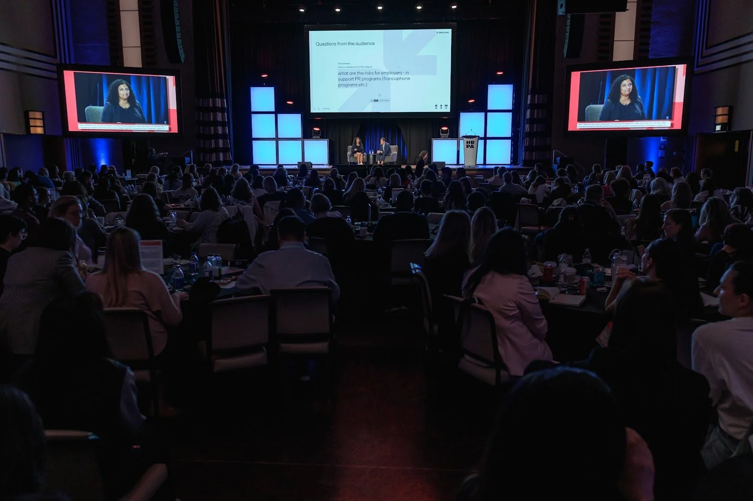 Large conference room filled with attendees sitting at tables, facing a stage with two speakers and large screens displaying a presentation.