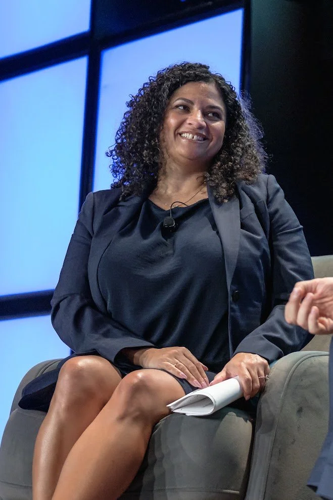 Woman with curly hair smiling, sitting on a gray chair, holding papers, wearing a black dress and blazer, against a blue-lit window background.