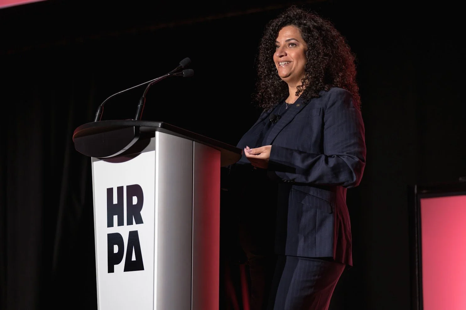 A woman in a dark pinstripe suit giving a speech at a podium with HRPA on it, on a black stage with red lighting.