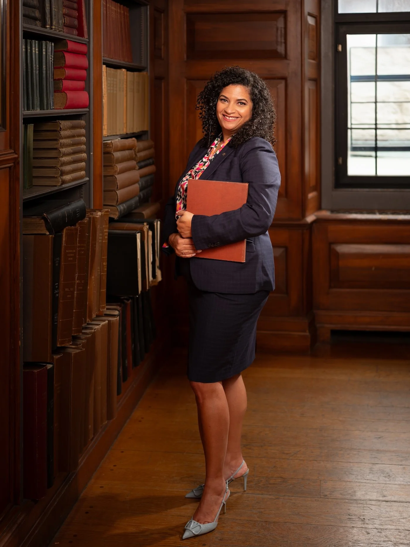 A woman with curly dark hair in a navy blazer and plaid skirt, holding a folder, standing in a wooden library near shelves of law books, smiling at the camera.