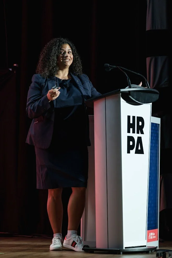 A woman with curly black hair, wearing a navy blazer, black dress, and white sneakers, stands behind a podium with 'HR PA' written on it, speaking at an event with dark curtains in the background.