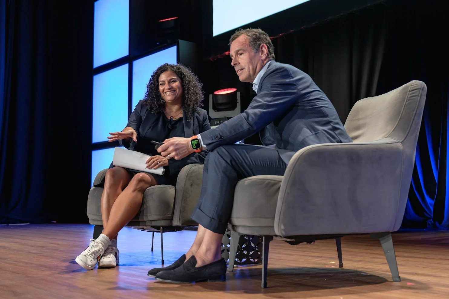 Two people sitting on stage in a discussion, with a woman smiling and a man holding a device, behind them is a large, bright screen.