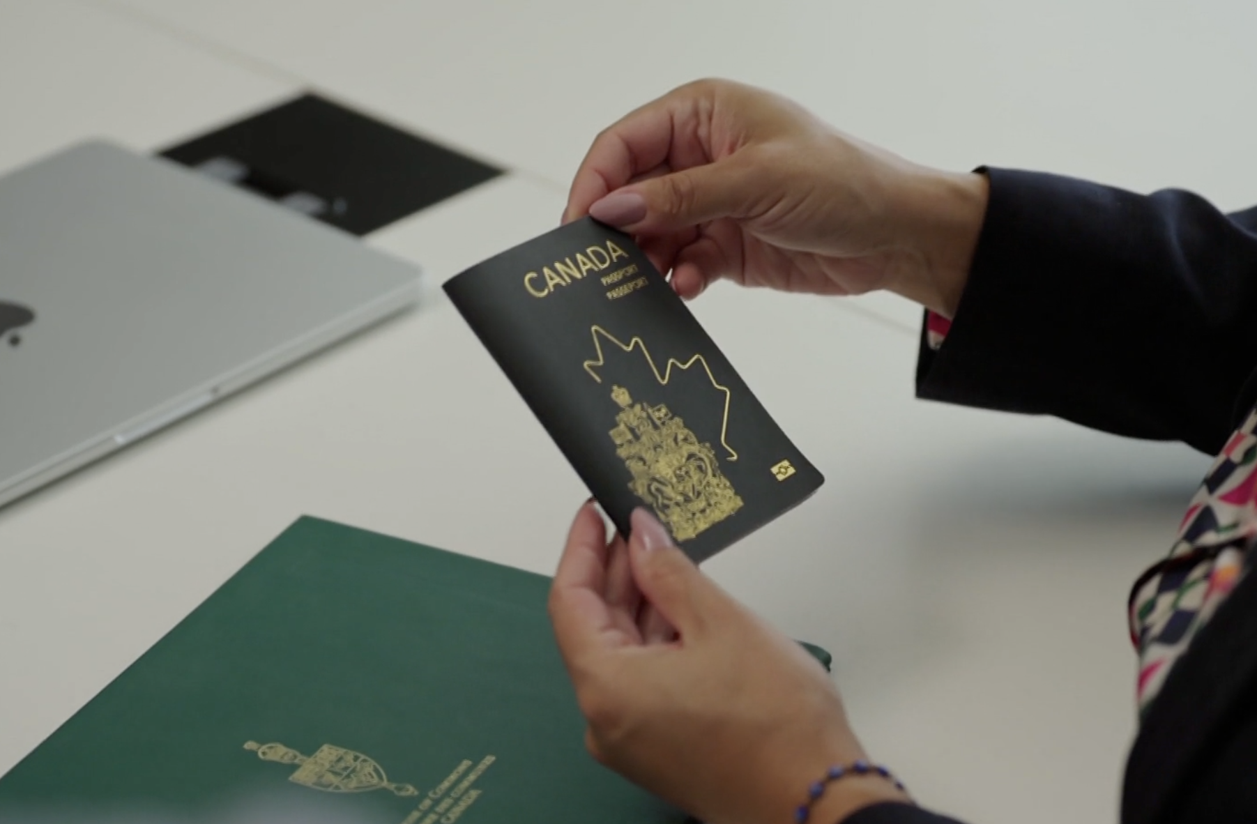 Close-up of a person holding a black and gold Canada passport with the image of a lion and a line drawing of the country on the cover, sitting at a white desk with a green folder and a silver laptop.